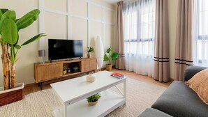 Een moderne woonkamer met een flatscreen televisie, een witte salontafel, een grijze bank en een groot raam met dunne gordijnen.