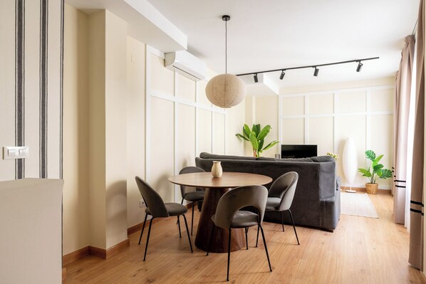 Una sala de estar moderna con una mesa redonda de madera, cuatro sillas, un sofá gris, un televisor y una lámpara colgante.