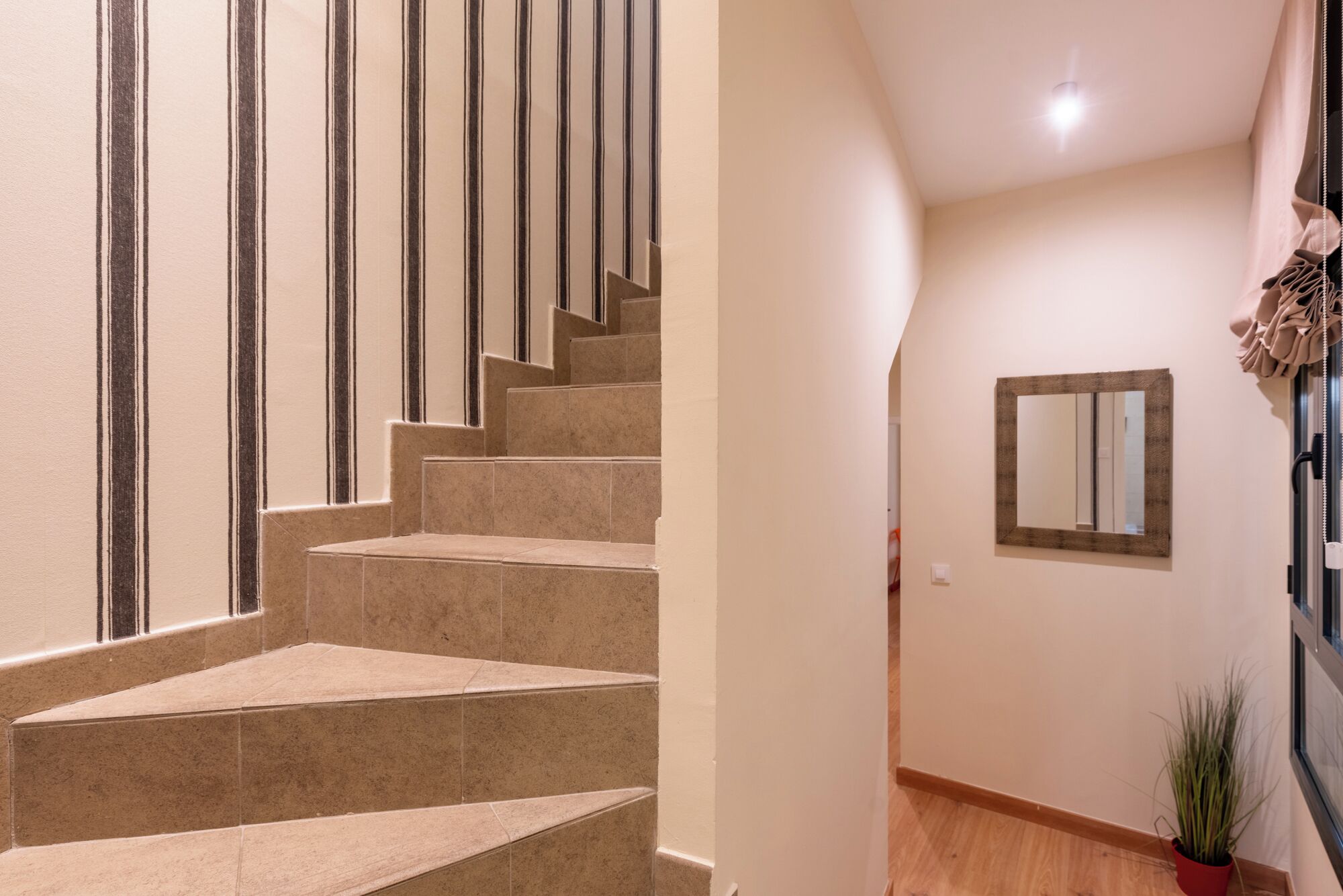 Un escalier recouvert de carreaux beiges et de bandes noires, menant à un palier agrémenté d’un miroir et d’une plante en pot.