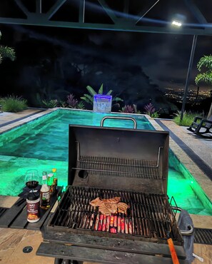 Ein Grill mit Essen, ein Schwimmbad und ein nächtlicher Blick auf eine Stadtlandschaft.