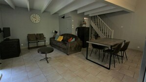 A living room with a sofa, a coffee table, a dining table with chairs, a clock on the wall, and a staircase leading to another level.