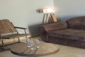 A round wooden table with three wine glasses, a wooden chair, and a brown sofa in a room with a tiled floor and a wall-mounted light fixture.