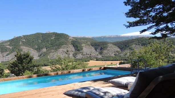 A wooden deck with a swimming pool, lounge chairs, and a view of green hills.