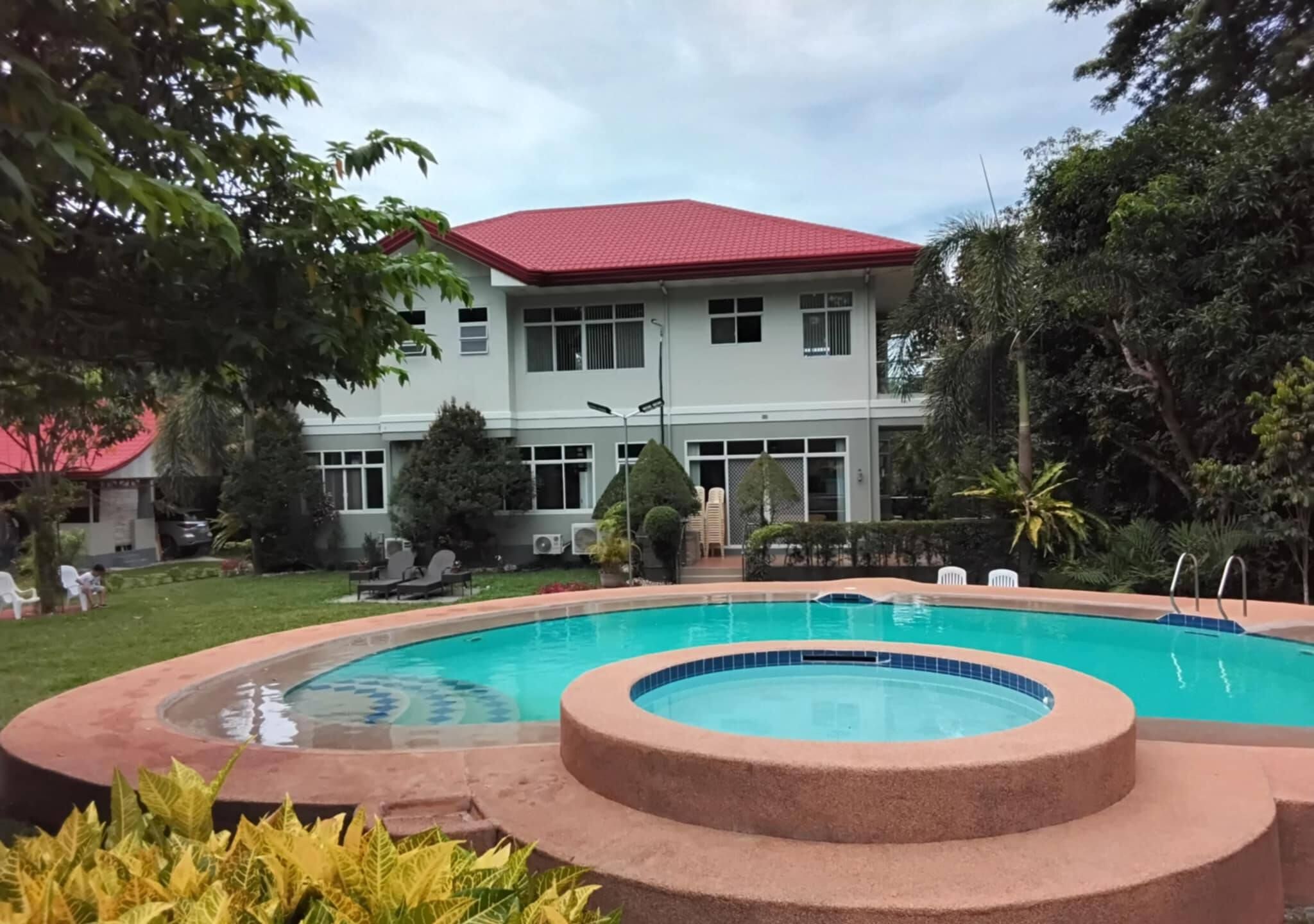 Una piscina con un jacuzzi circular, rodeado de un jardín ajardinado y un edificio de techo rojo al fondo.
