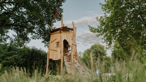A rustic wooden structure with a ladder leading up to a small door, surrounded by lush greenery. - Luxury Family Tent with Play Area (Balkbrug)