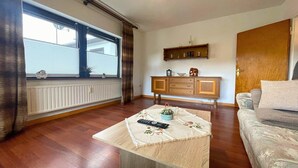 Ein Wohnzimmer mit Holzboden, einer Couch, einem Tisch mit Fernbedienung, einem Fenster mit Vorhängen, einem Schrank und einer Tür.