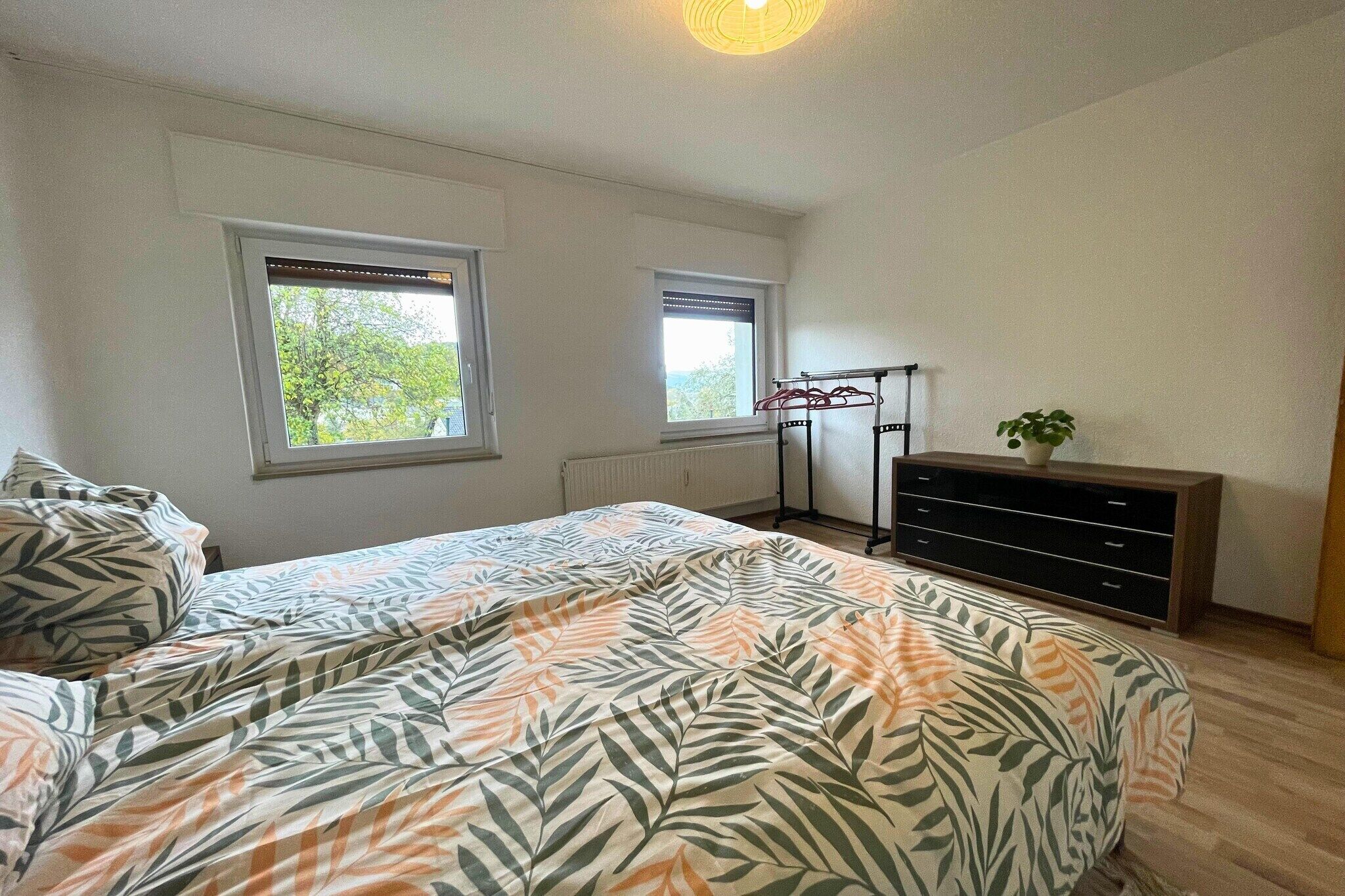 Een slaapkamer met twee ramen, een bed met een bladprintdeken, een radiator en een dressoir met een plant.
