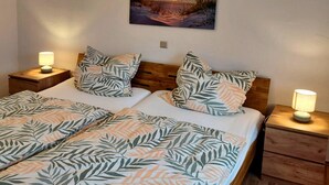 A double bed with leaf-patterned bedding, two bedside tables with lamps, and a sunset painting on the wall. - Holiday Home in Birresborn with balcony (Birresborn)