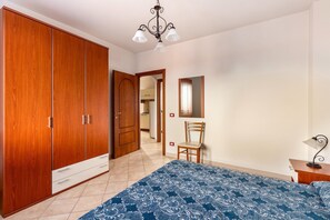 A bedroom with a large wardrobe, a bed with a patterned bedspread, a chair, a desk with a lamp, and a mirror on the wall. - 150 meters from the sea, 2 swimming pools (Valledoria)