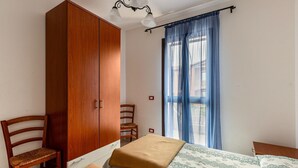 A bedroom with a wooden wardrobe, a wooden chair, a bed with a patterned bedspread, and a window with blue curtains. - 150 meters from the sea, 2 swimming pools (Valledoria)
