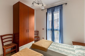 Ein Schlafzimmer mit einem Holzschrank, einem Einzelbett mit gemustertem Bettbezug, einem Holzstuhl und einem Fenster mit blauen Vorhängen.