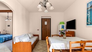 A room with a wooden sofa, a dining table with chairs, a TV on the wall, and a door with a sign. - 150 meters from the sea, 2 swimming pools (Valledoria)