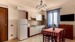 Eine kompakte Küche mit Kühlschrank, Herd und gefliestem Fliesenspiegel. Ein Esstisch mit Stühlen und ein an der Wand befestigtes Fernsehgerät.