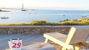 Terraza de madera con un banco y una bolsa marcada con el número 29, con vista a un mar tranquilo con barcos y un faro a lo lejos.