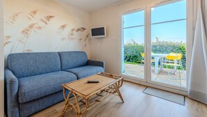 Ein Zimmer mit blauer Couch, einem Holztisch, einem an der Wand befestigten Fernseher und einer Glastür, die zu einem Außenbereich mit gelben Stühlen führt.