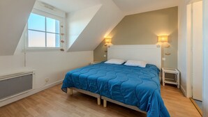 Ein Schlafzimmer mit schrägem Dach, einem großen Fenster, einem Bett mit blauer Bettdecke, zwei Nachttischlampen und einer Heizung.