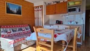 Une pièce chaleureuse avec un mur en bois, un canapé recouvert d’une couverture aux motifs festifs, une table à manger avec des chaises, un coin cuisine doté de placards et d’appareils électroménagers, et une fenêtre donnant sur les arbres.