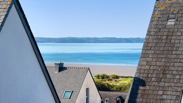 Une vue sur la côte, prise depuis un bâtiment doté d’un toit en pente, donnant sur une plage et la mer.