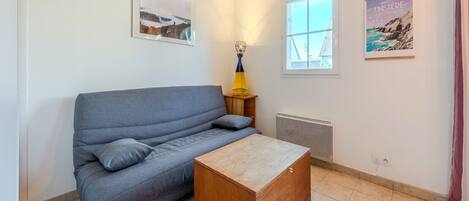 Une pièce avec un canapé gris, une table d’appoint en bois sur laquelle se trouve une lampe, un radiateur et deux tableaux encadrés accrochés aux murs.