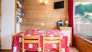 A room with a wooden ceiling, a bed with a red polka dot bedspread, a small table with a radio, and a window with a red curtain.