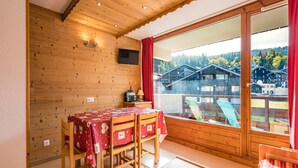 A wooden cabin with a dining area, a TV mounted on the wall, and a large window offering a view of buildings and trees.