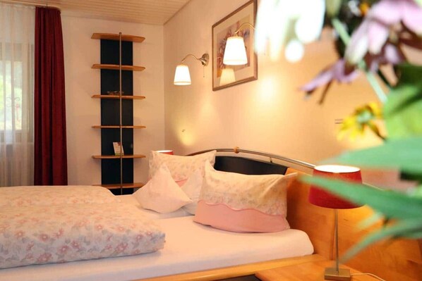 Habitación de hotel con cama, mesita de noche, lámpara, estante y ventana con cortinas.