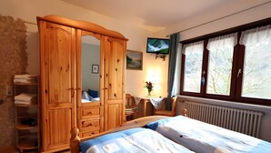 Een slaapkamer met een houten garderobe, een bed met een bedovertrek met patroon, een televisie, een raam met gordijnen en een radiator.
