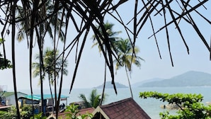 A view of a beach with palm trees, a small building with a red roof, and a distant mountain range. - Grand Sea Shades Goa (Canacona)
