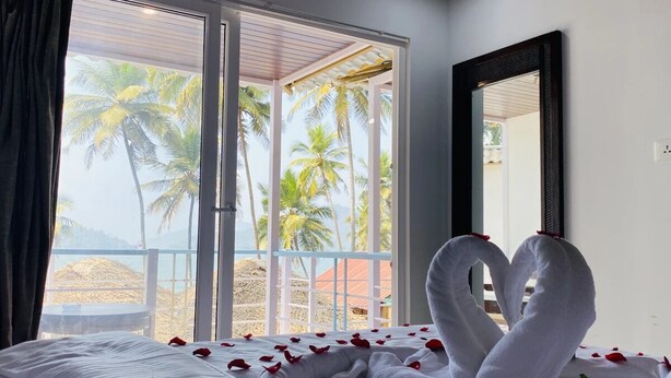A hotel room with a bed, a large window with a view of palm trees and a beach, and a heart-shaped towel arrangement. - Grand Sea Shades Goa (Canacona)