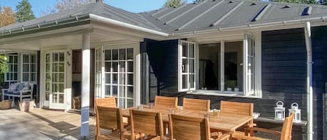 En overdækket terrasse med træmøbler og udsigt til huset.