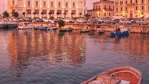 A waterfront scene with a rowboat filled with fishing nets and equipment docked in the foreground. In the background, there is a large, classical building with multiple windows and a balcony. - A stone's throw from the cathedral: authentic Sicilian house in Ortigia (Siracusa)