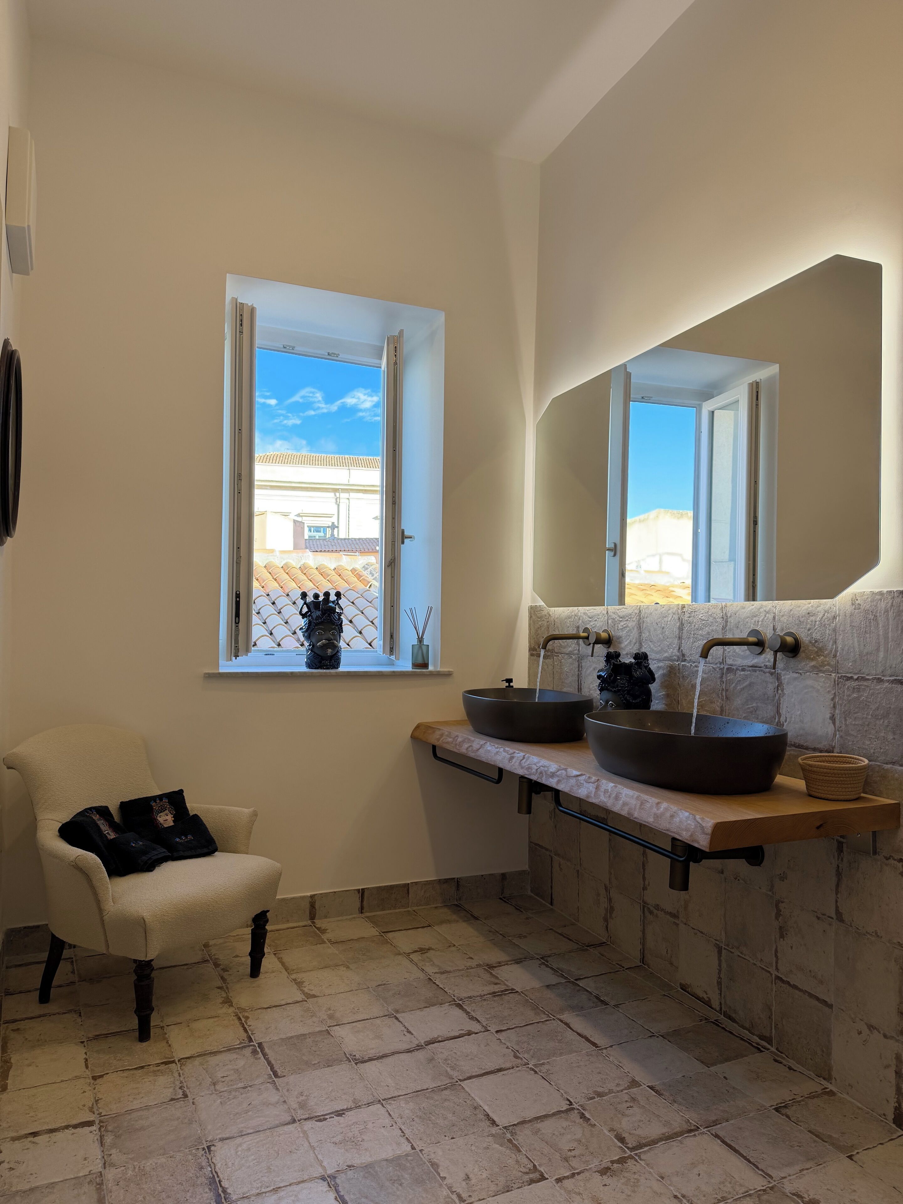 A bathroom with a large window, a white chair, a sink with a vessel basin, and a stone wall.