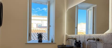 A bathroom with a large window, a white chair, a sink with a vessel basin, and a stone wall.