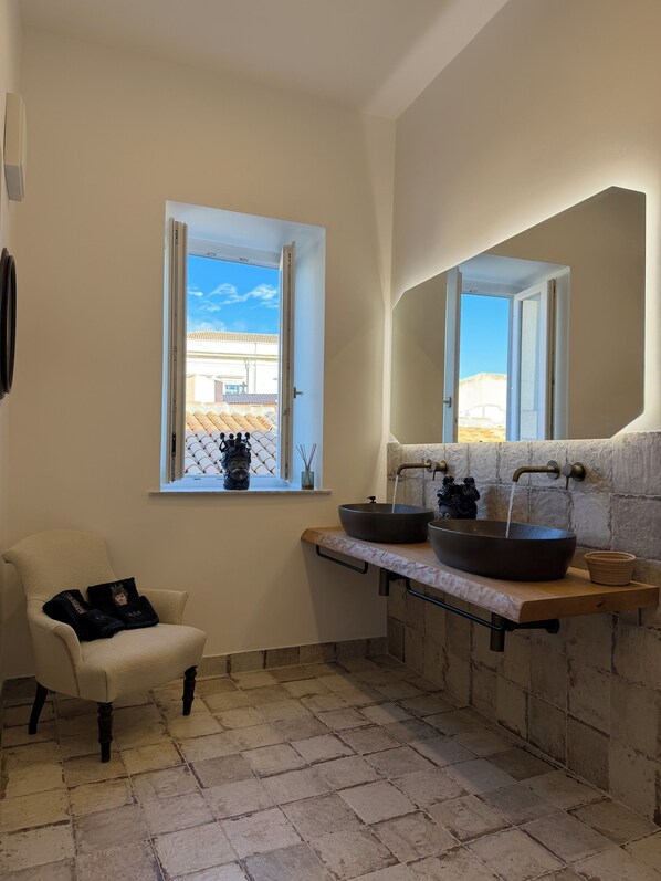 Un baño con un ventanal grande, una silla blanca, un lavabo con lavatrastos y una pared de piedra.