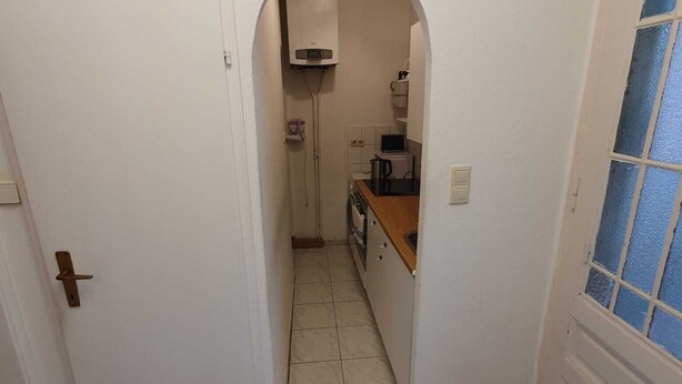 Un pasillo estrecho con una puerta blanca a la izquierda y una ventana con marco azul a la derecha. Se puede ver una zona de cocina a través de un arco en el centro, que incluye una estufa, un fregadero y una encimera.