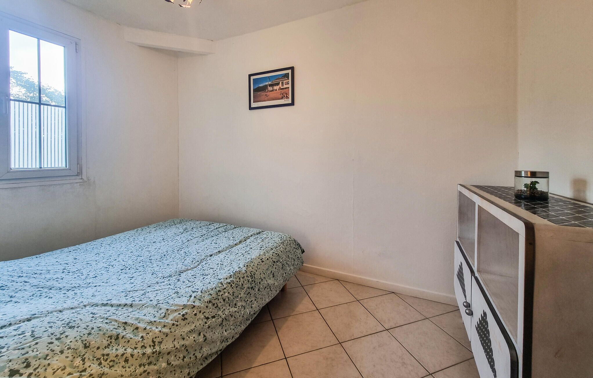 A small, sparsely furnished room with a bed, a small cabinet, and a window.