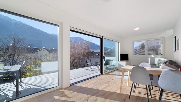 A modern interior with a large glass door leading to an outdoor deck, a dining area with chairs, and a flat-screen TV on a stand. - Apartment 'Naturns Mountain Suite' with Mountain View, Private Terrace and Wi-Fi (Naturno Naturns BZ)