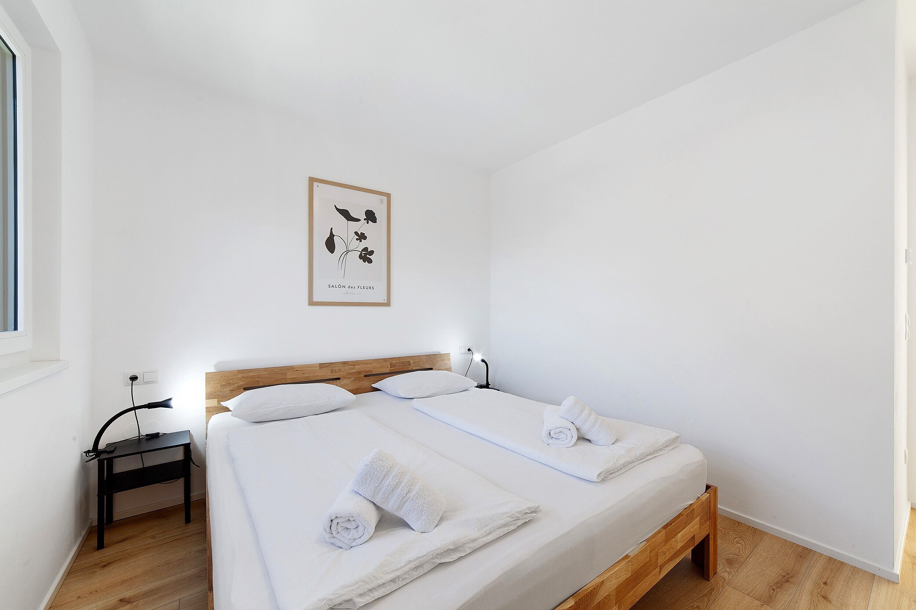 Een nette, minimalistische hotelkamer met een houten bed en witte beddengoed, plus een klein bureau met een lamp.