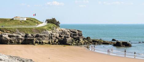 A coastal landscape with a sandy beach, rocky cliffs, and a flag on a hill.