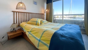 Una cama con cabecera de madera, colcha a rayas azul y amarilla, y manta azul. Dos lámparas fijadas a la pared con diseño tejido. Una mesita de noche con lámpara al lado de la cama.