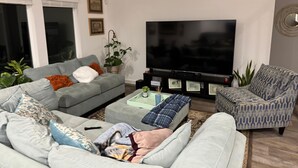 Ein Wohnzimmer mit grauem Sofa, einem gemusterten Sessel, einem Couchtisch und einem Flachbildfernseher.