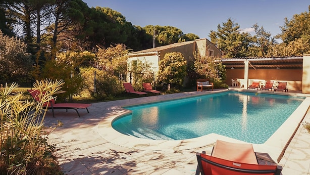 A swimming pool with lounge chairs and a covered area, surrounded by trees and a building.