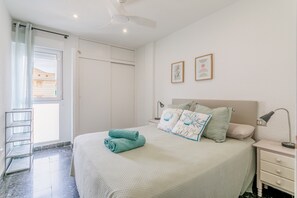 A bedroom with a bed, a nightstand, a lamp, a ceiling fan, a window with a curtain, and a door.