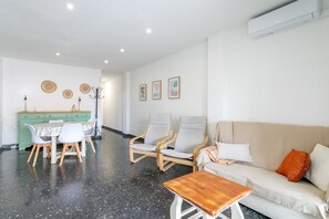 A modern living room with a sofa, chairs, a wooden coffee table, a dining table with chairs, and a kitchen area in the background.