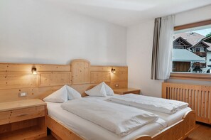 Une chambre avec une tête de lit en bois, deux lits avec du linge de lit blanc, une table de chevet en bois dotée d’un tiroir et une fenêtre donnant sur des immeubles.