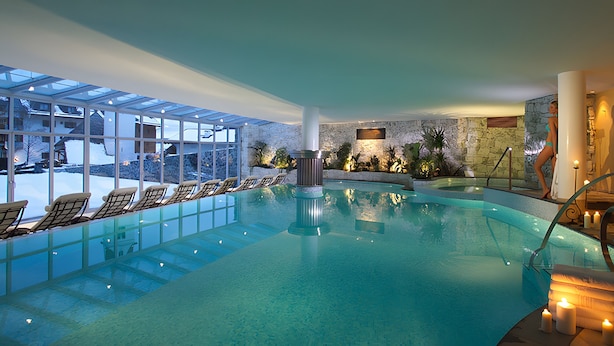 Una piscina interior con paredes de vidrio, sillas de descanso y una pared de piedra con plantas.