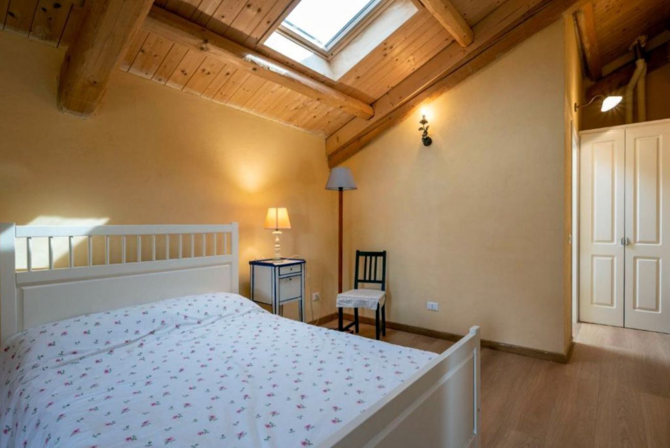 Een slaapkamer met een houten plafond, een bed met een bloemenprint dekbedovertrek, een nachtkastje met een lamp, een stoel en een kledingkast.