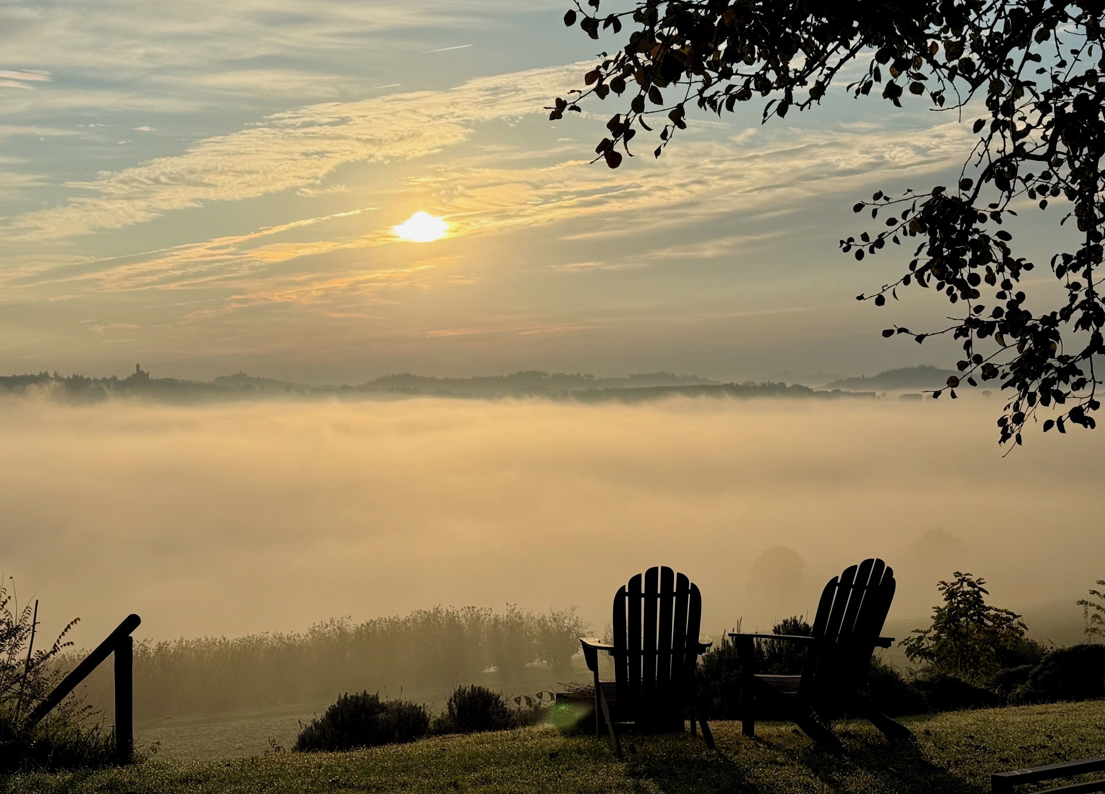 A sunrise over a foggy landscape with two Adirondack chairs facing a body of water.