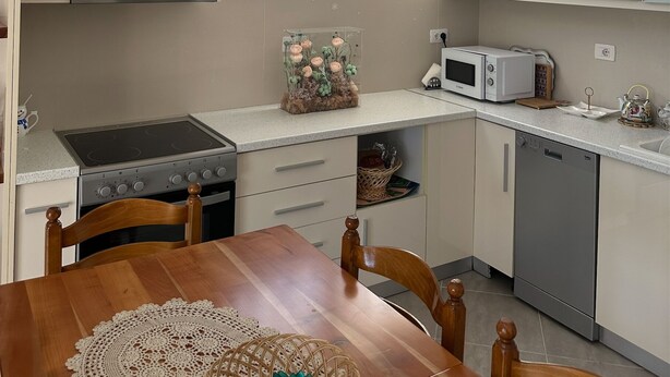 A compact kitchen with white cabinets, a microwave, a stove, and a dishwasher. There is a wooden dining table with chairs, a decorative vase, and a clear glass container with flowers. - Olive Hill Apartment (Farkë)