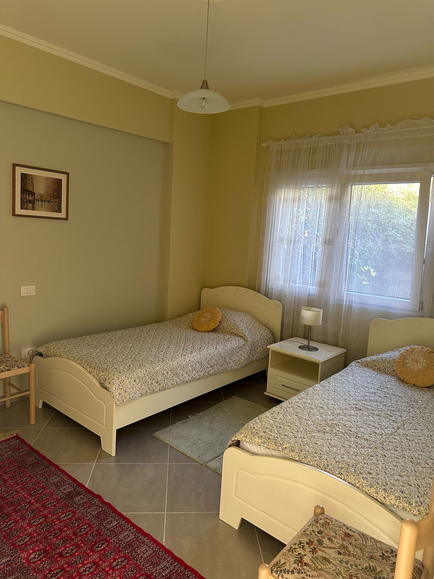 Une chambre avec deux lits simples, une table de chevet avec une lampe, une chaise, un tapis rouge à motifs et une fenêtre avec des rideaux légers.